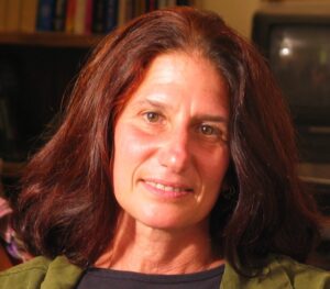 A person with shoulder-length brown hair and a warm smile is looking at the camera. They are wearing a dark shirt and a green jacket. The background is blurred but contains some shelves with books, hinting at their involvement in the Cal NORML Board of Directors, and a television screen. CA Norml