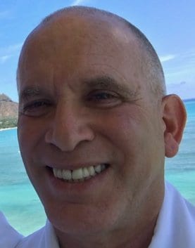 A close-up photo of a man smiling at the camera with a scenic background of the ocean and blue sky. He is wearing a white shirt and appears to be outdoors, embodying the relaxed spirit of Cal NORML's Board of Directors. CA Norml