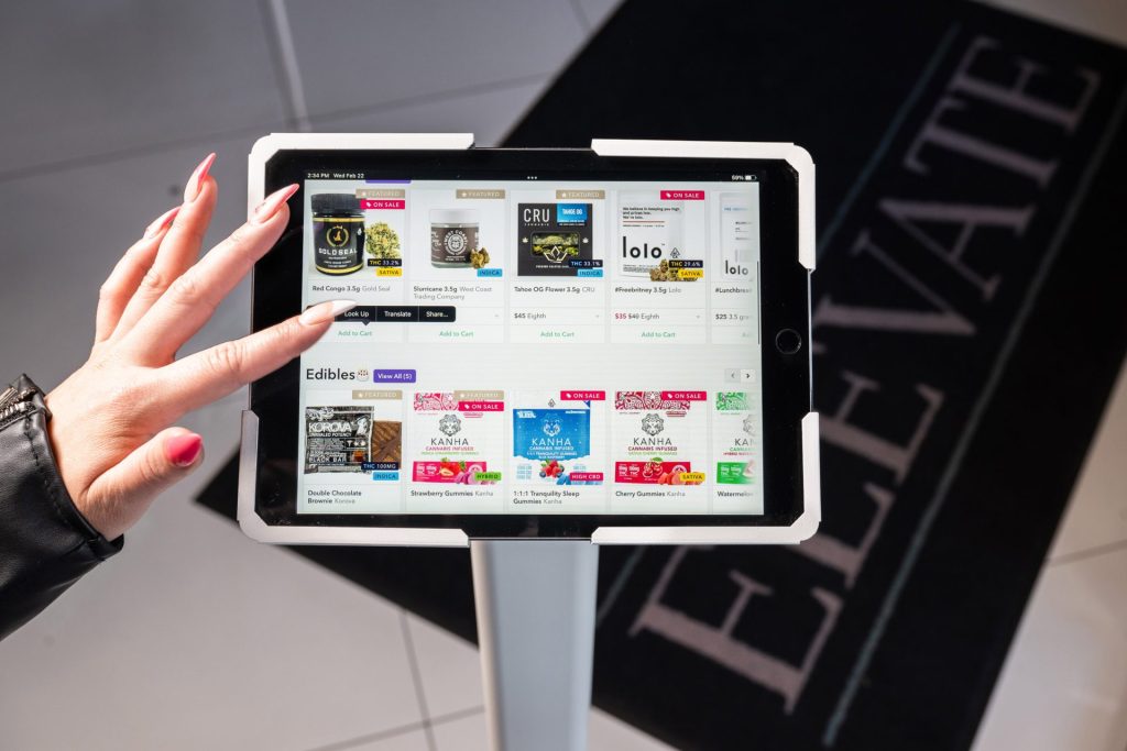 A person uses a touchscreen tablet displaying a selection of cannabis products; part of a black mat with the word "ELEVATE" is visible on the floor. Ca NORML