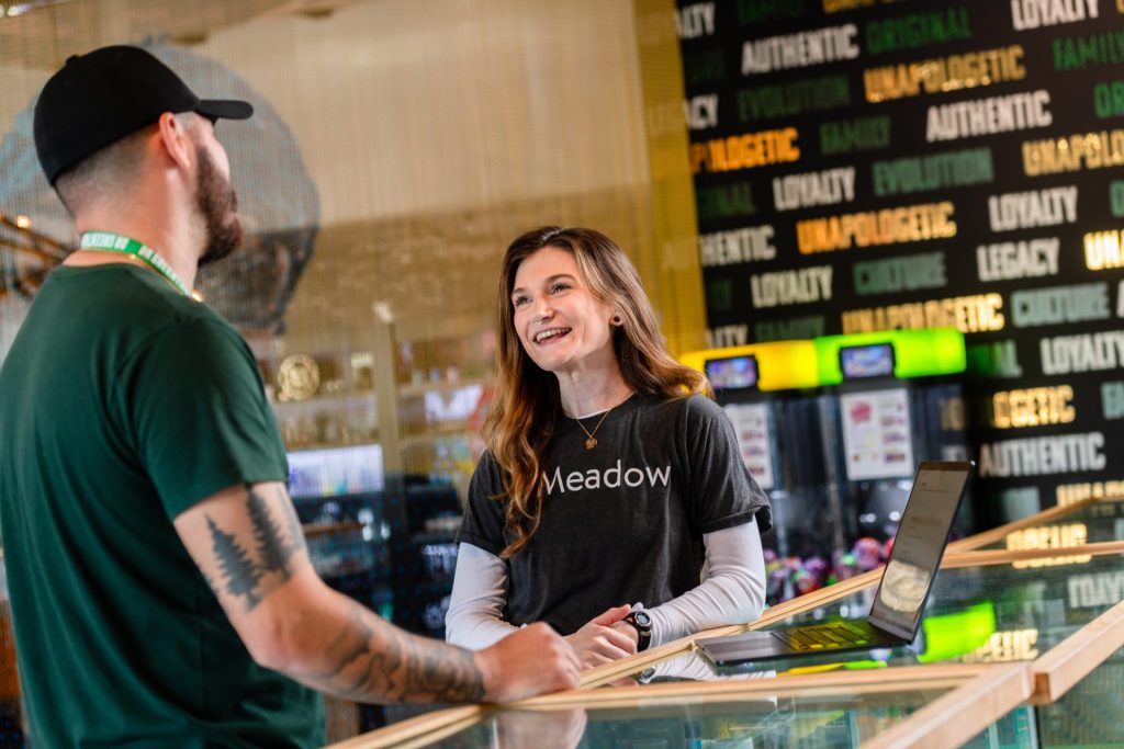 A woman in a "Meadow" shirt smiles while talking to a man at a counter. A laptop and display case are in front of her. The background features words like "AUTHENTIC" and "LOYALTY" on the wall. Ca NORML