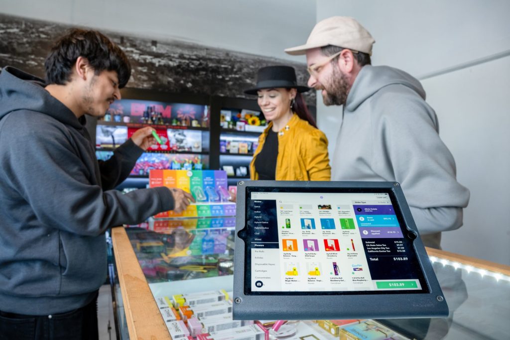 Three people stand at a glass counter in a store with colorful products. A point-of-sale screen in the foreground shows transaction details and items for sale. Ca NORML
