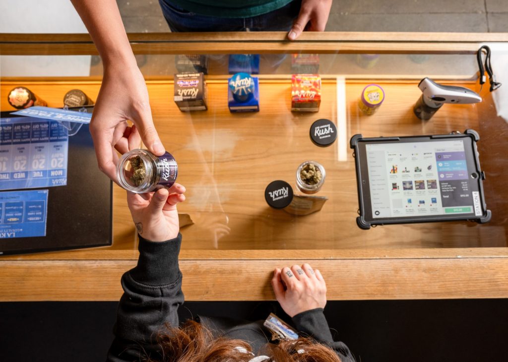 A person behind a counter hands a clear jar with cannabis buds to a customer. Cannabis containers and a tablet displaying a sales screen are on the countertop. Ca NORML