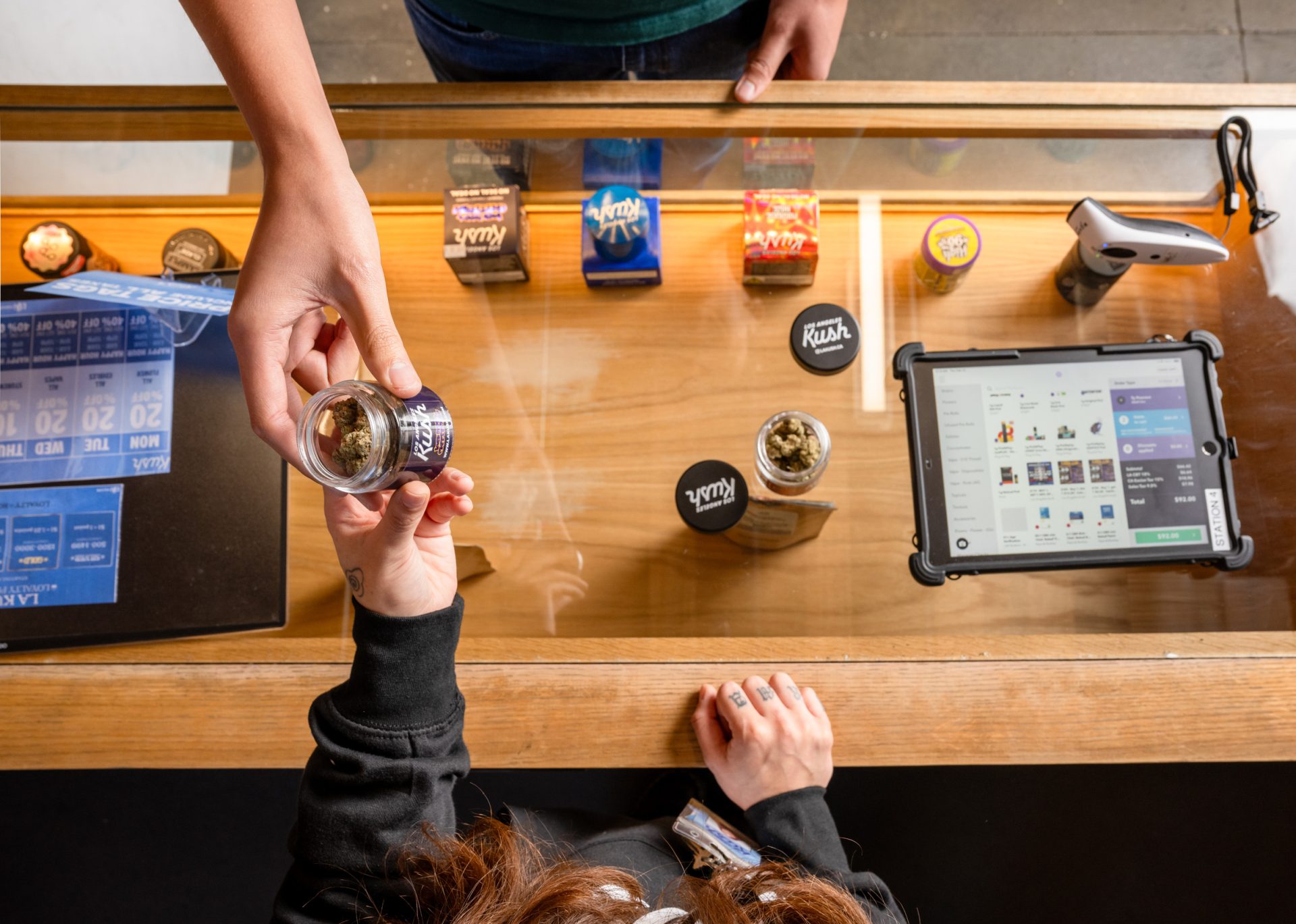 A person behind a counter hands a clear jar with cannabis buds to a customer. Cannabis containers and a tablet displaying a sales screen are on the countertop. Ca NORML