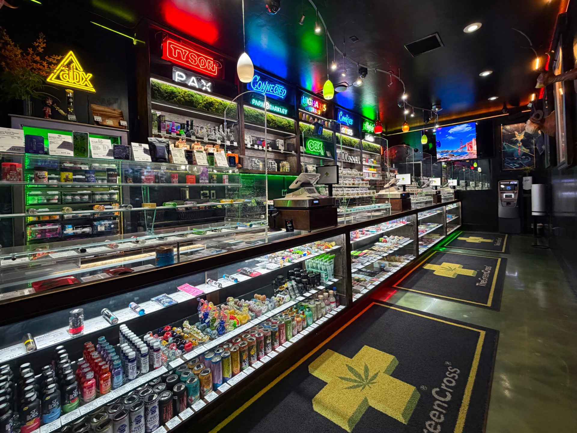 A brightly lit dispensary, The Green Cross, features glass display cases with various cannabis products, drinks, and accessories. Neon signs for brands line the walls above, and branded floor mats are visible on the glossy floor. Ca NORML