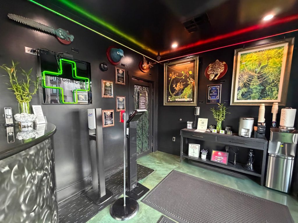 A modern dispensary lobby with green and red LED lights, a prominent Green Cross sign, framed art on black walls, a counter adorned with bamboo plants, a coffee station, and clear informational displays. Ca NORML