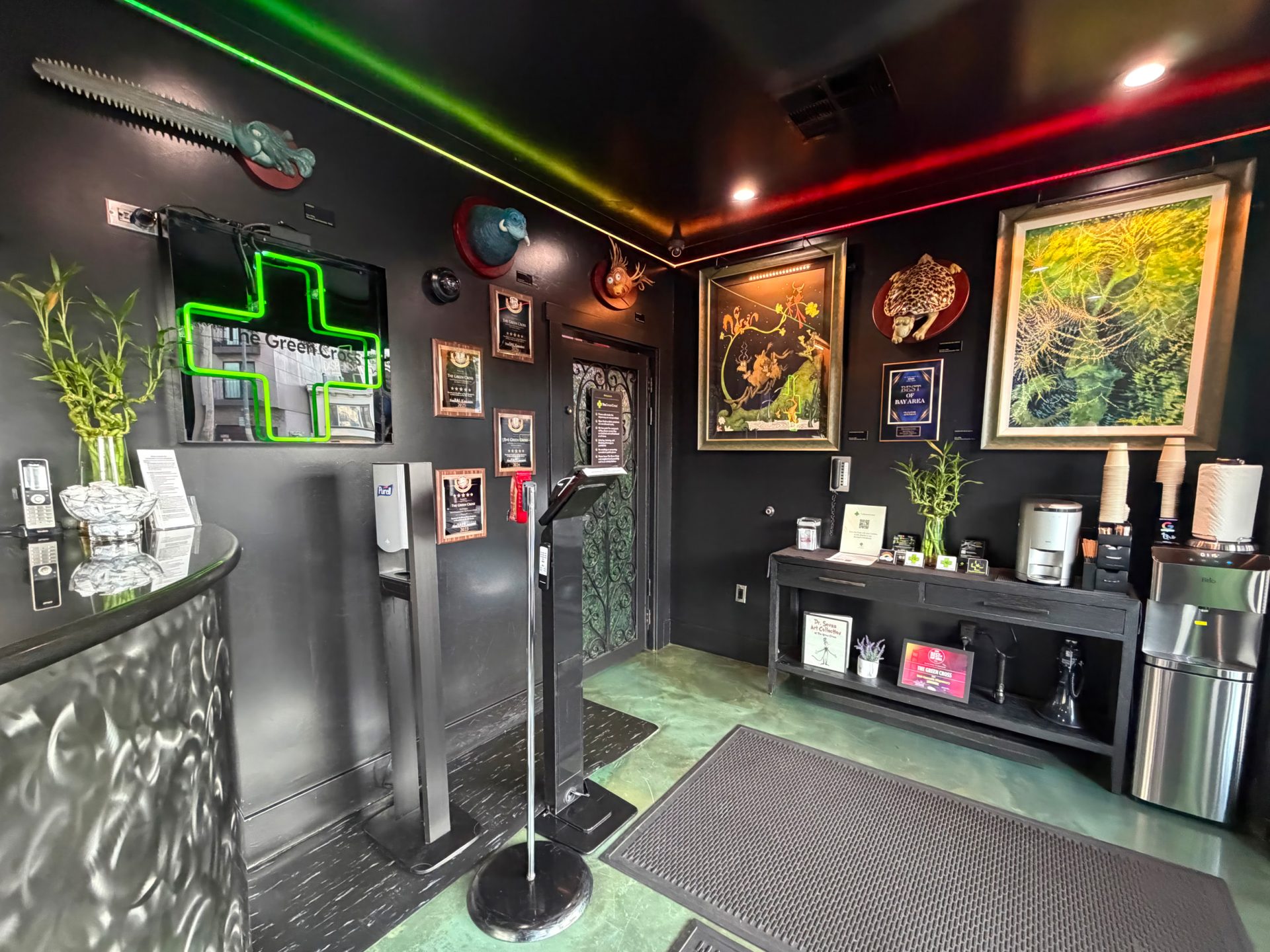 A modern dispensary lobby with green and red LED lights, a prominent Green Cross sign, framed art on black walls, a counter adorned with bamboo plants, a coffee station, and clear informational displays. Ca NORML