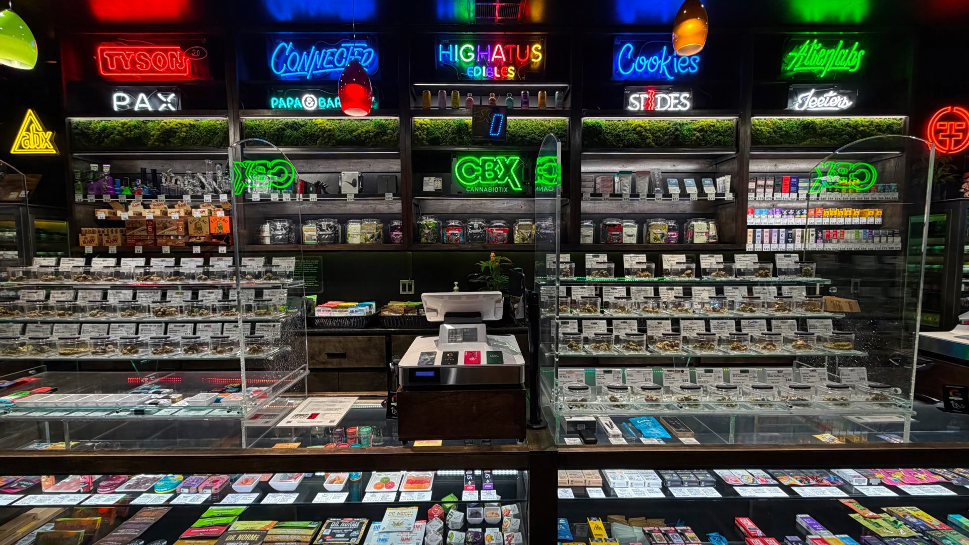 A well-lit cannabis dispensary counter at The Green Cross displays glass jars of marijuana, vape cartridges, edibles, and various products. Neon signs above shelves advertise brands like Highatus, Cookies, and CBX. A cash register sits at the center. Ca NORML