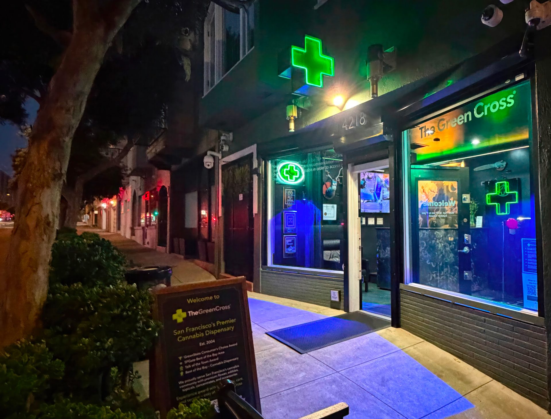 A brightly lit cannabis dispensary called The Green Cross, with glowing green neon crosses above the entrance, is open at night on a city street. A sidewalk sign welcomes visitors near the doorway to Green Cross’s inviting atmosphere. Ca NORML