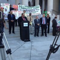 A group of people stand on the steps of a building with columns, holding signs advocating for marijuana decriminalization in California. A man speaks at a podium with cameras set up in the foreground to record the event. The signs include messages like "DE-CRIMINALIZE CANNABIS HEMP. CA Norml