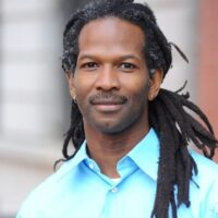 A person with long dreadlocks and a mustache, reminiscent of Drs. Carl Hart's style, is wearing a light blue button-up shirt. They are standing outdoors in front of a blurred building facade with a neutral expression and small hoop earrings in both ears. CA Norml
