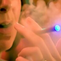 A close-up of a person inhaling from an e-cigarette, with visible vapor and a blue light at the tip of the device. The person's mouth is slightly open, and the image is slightly blurred due to the vapor, highlighting how policies like E-CIG BILL SB 140 aim to regulate such devices in non-smoking areas. CA Norml
