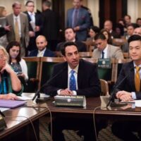 A group of people sit at a table in a busy room, likely a governmental or corporate meeting. Three individuals are in the foreground: a woman on the left, a man in the center speaking about the DUID Bill, and a man on the right. People in professional attire are seated and standing in the background. CA Norml