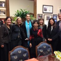 A group of eight reformers, dressed in formal business attire, stand in an office setting on Capitol Hill, smiling at the camera. The room has framed certificates and photographs on the walls, a green plant on a wooden cabinet, and a table with floral arrangements in the foreground. CA Norml