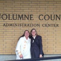 Two women standing and smiling in front of the Tuolumne County Administration Center. The woman on the left is wearing a white garment, and the woman on the right is wearing a dark jacket and glasses. They appear happy and are posing closely together, perhaps discussing the recent dispensary ban in Tuolumne County. CA Norml