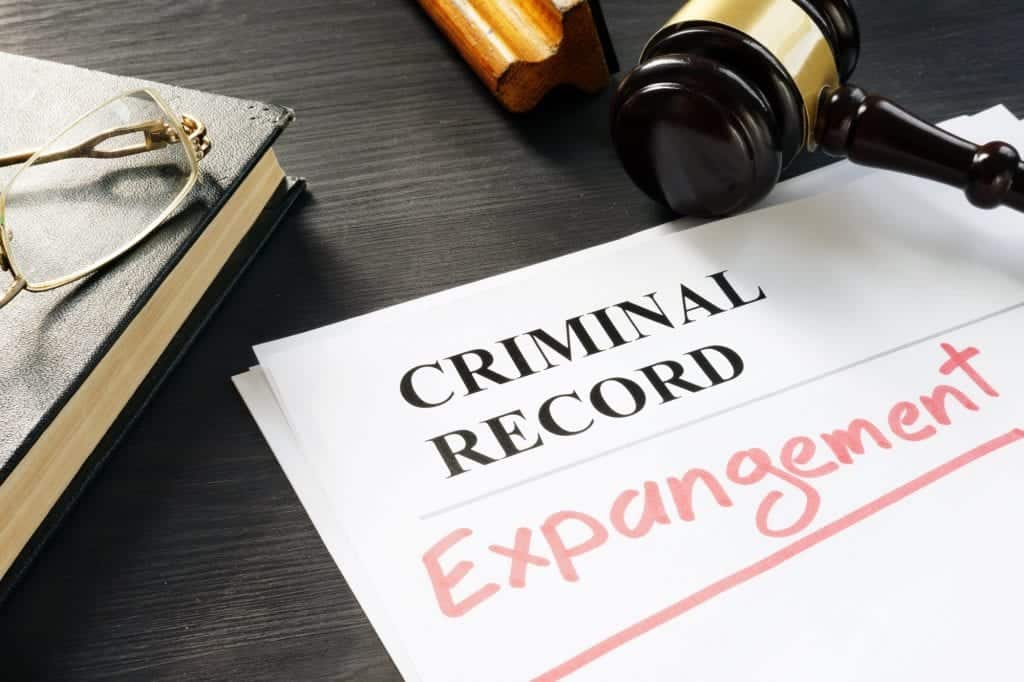 A document labeled "CRIMINAL RECORD" with the word "Expungement" written in red sits on a dark desk. Nearby are a pair of glasses, a closed book, and a judge's gavel. The scene signifies steps taken in California for clearing past marijuana crimes from individual records. CA Norml