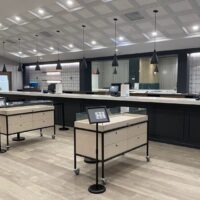 A modern, spacious retail store interior with a long counter lined with pendant lights. Four display tables on wheels are in the foreground, each holding framed signs. Reminiscent of trendy California cannabis businesses, the store features a stylish black and white decor with a partial tiled wall. CA Norml