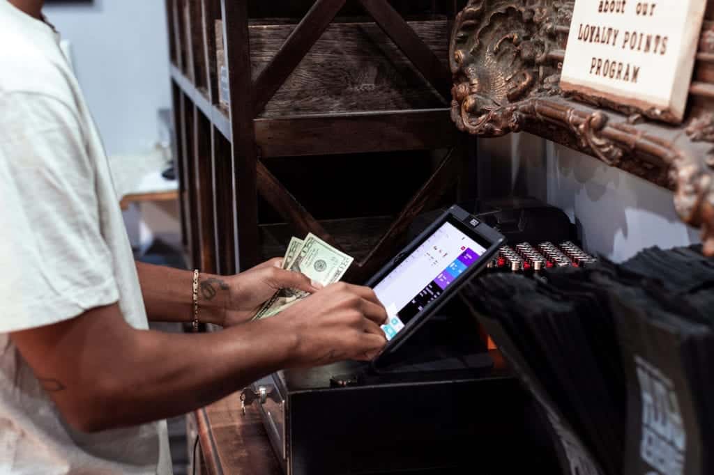 A person is standing at a cash register, holding a few dollar bills in one hand and operating a touch screen with the other. In the background, there is a sign for a loyalty points program and a shelf with various items promoting local businesses. CA Norml