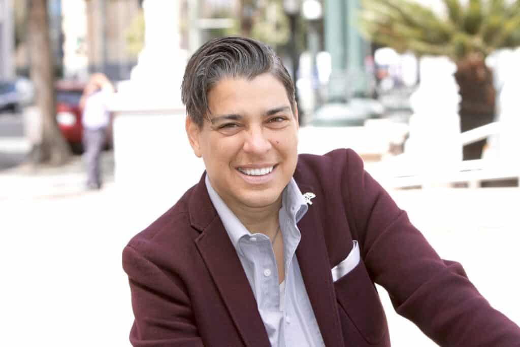 A smiling person with short dark hair, dressed in a maroon blazer and light-colored shirt, poses for a photo outdoors. The background features a street scene with blurred trees, buildings, and people after attending an Oakland Public Safety Committee meeting. CA Norml
