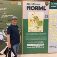 A man stands smiling next to a large California NORML poster at an event on July 25, 2023. The poster features a woman holding a flag and a marijuana leaf, offering information about Cal NORML’s advocacy for marijuana law reform. The man is wearing a cap and holding a bag. CA Norml