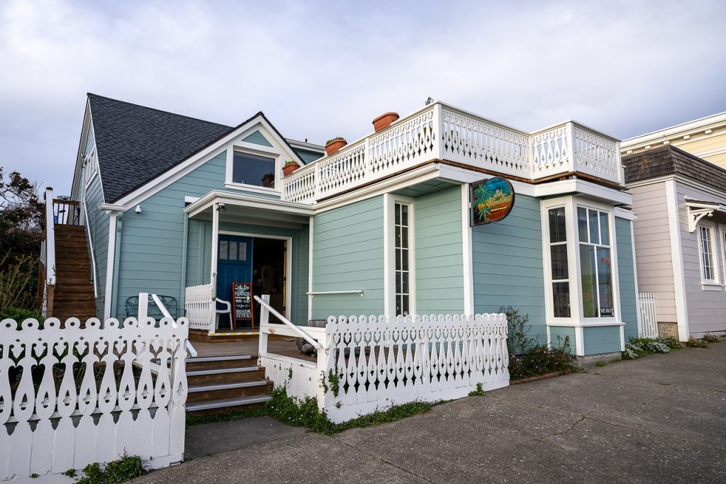 A charming blue, two-story building with white trim, complete with a decorative white picket fence and a wooden deck. Potted plants adorn the rooftop, next to a colorful sign advertising Organic and Local Cannabis. The surroundings include a cloudy sky and neighboring buildings. CA Norml