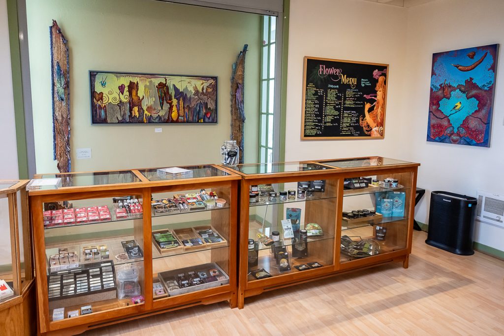 A brightly lit store with wooden display cases showcasing various products, including Sungrown Organic and Local Cannabis. Behind the cases, there's a colorful "Flower Menu" board listing items and prices. The walls feature vibrant artwork, adding a creative ambiance to the space. CA Norml