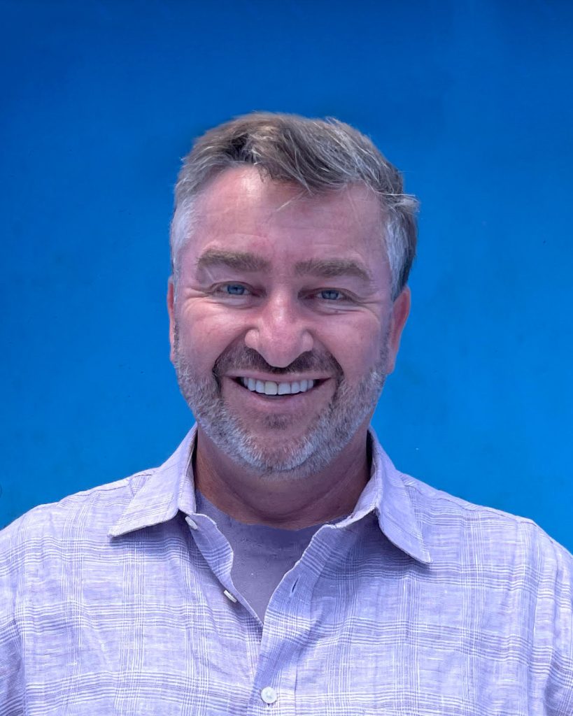 Ian Rassman, a middle-aged man with short, light brown hair and a beard, smiles in front of a bright blue background. He is wearing a light purple collared shirt over a lilac T-shirt. Ca NORML