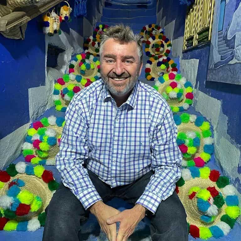 Ian Rassman, a man with a beard and short hair, is sitting on colorful pom-pom decorated stairs. He is wearing a blue and white checkered shirt and smiling towards the camera. The background is vibrant and festive with a blue theme and multiple colorful hats arranged on the steps. CA Norml