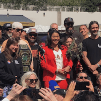 A diverse group of people, including Mayor Breed, smile and pose for a photo outdoors. Some hold a large belt that appears to be an award. The backdrop showcases greenery, a white wall, and a small structure, all under the sunny skies of SF Weed Week. CA Norml