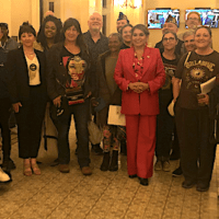 A diverse group of people stand in a lobby, some holding papers. At the center, a woman in a red pantsuit smiles. One person uses a wheelchair. The environment appears formal, with chandeliers and carpeted flooring, suggesting an official or celebratory event perhaps related to Compassion Programs. CA Norml