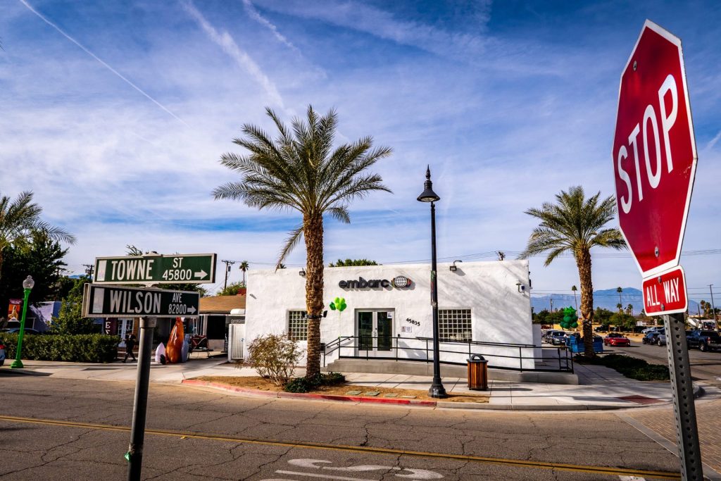 Exterior view of the Embarc dispensary in Indio, California, set against a sunny backdrop with palm trees, featuring a white building and the Embarc sign, near the intersection of Towne Street and Wilson Avenue.