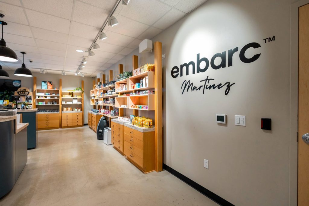 Interior view of the Embarc dispensary in Martinez, California, displaying neatly organized wooden shelves filled with cannabis products, with the Embarc Martinez logo on the wall.