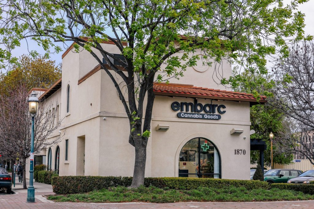 Exterior view of the Embarc dispensary in Redwood City, California, showcasing a tan-colored building with a tiled roof, surrounded by trees, with the Embarc sign on the facade.