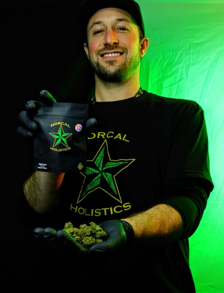 Male wearing Norcal holistics branded tshirt holding cannabis flower in one hand and bags of flower in another.