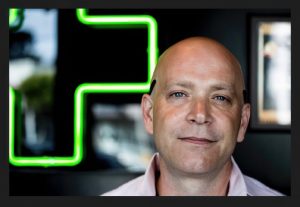 A bald man with blue eyes and a slight smile stands indoors, with a bright green neon cross in the background and framed art on a dark wall, as if ready to share testimonials often collected by retailers. CA Norml