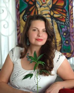 A woman with long brown hair, wearing a white blouse and turquoise jewelry, sits indoors holding a green cannabis plant. Behind her is a colorful tapestry featuring an elephant and butterfly wings—often seen in retailers’ testimonials. CA Norml