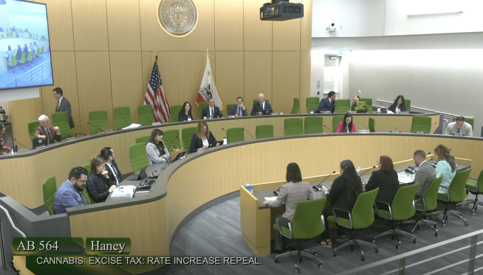A government meeting in a large, modern chamber with people seated in a semicircle. Two American flags and one California flag are displayed. A screen reads “AB 564 Haney Bill on Cannabis Tax Increase Repeal,” backed by Cal NORML. CA Norml