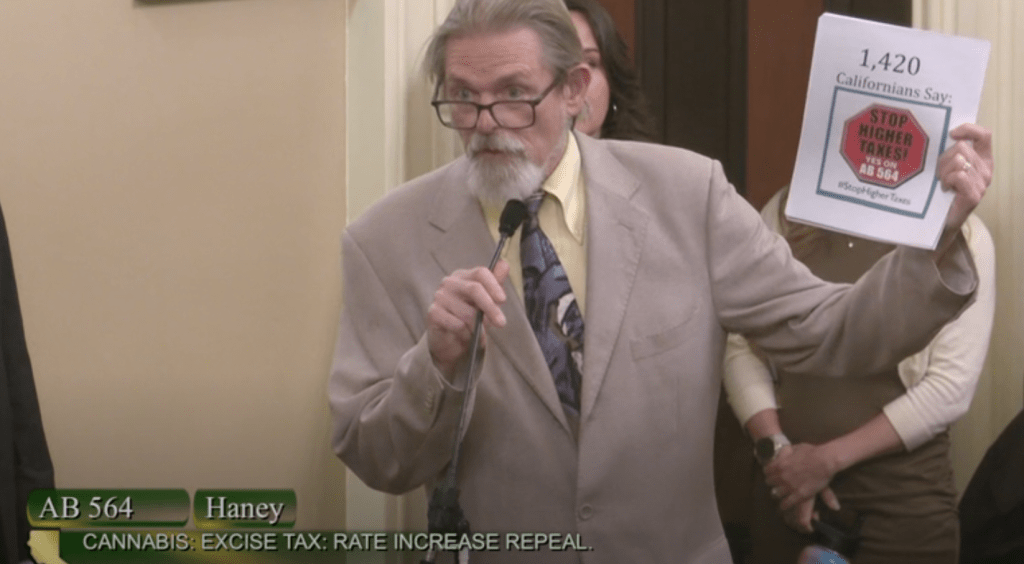 An older man in a beige suit speaks into a microphone while holding a paper that reads "1,420 Californians Say: STOP HIGHER TAXES!" at a California legislative session discussing the cannabis industry and tax increase repeal (AB 564). CA Norml