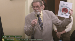 An older man in a beige suit speaks into a microphone while holding a paper that reads "1,420 Californians Say: STOP HIGHER TAXES!" at a California legislative session discussing the cannabis industry and tax increase repeal (AB 564). CA Norml