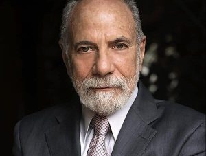 An older man with a neatly trimmed white beard and short hair, wearing a gray suit, white shirt, and patterned tie, looks confidently at the camera against a dark background. CA Norml