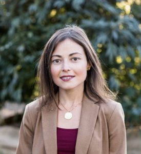 Lauren Mendelsohn, a woman with straight brown hair, wearing a tan blazer over a maroon top and gold necklace, stands outdoors with blurred green foliage in the background. She is looking at the camera and smiling softly. CA Norml