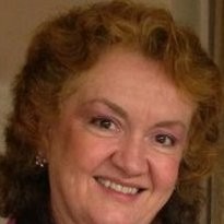 Jackie Wilson, a woman with curly, reddish-brown hair, smiles warmly at the camera. She is wearing light makeup and earrings, and the background is softly blurred. CA Norml