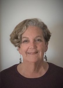 Denise Martellacci, an older woman with short, wavy gray hair, smiles gently at the camera. She is wearing dangling earrings and a dark top, standing against a plain, light-colored background. CA Norml