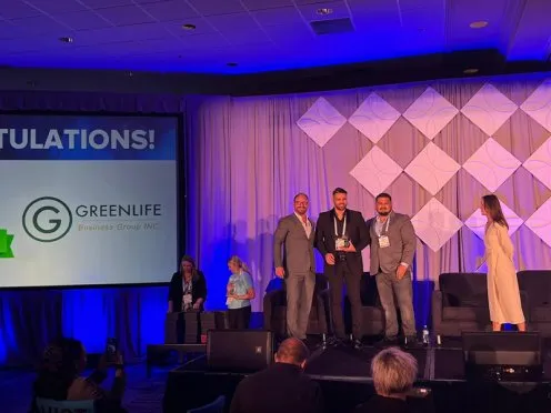 Four men in suits stand on stage receiving an award for Green Life Business, renowned cannabis business brokers, while two women look on. The screen displays "CONGRATULATIONS!" and the GreenLife logo, with blue lighting and a geometric wall pattern in the background. Ca NORML