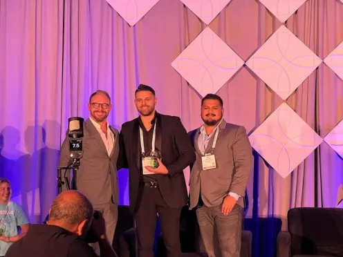 Three men in suits stand on a stage, one holding an award, photographed as a woman sits to the side. The backdrop features geometric panels and purple lighting—an event hosted by Green Life Business Group, a leader in cannabis business brokerage. Ca NORML
