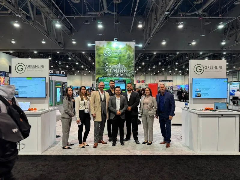 A group of eight people in business attire smile at a trade show booth for Green Life Business, cannabis business brokers specializing in businesses for sale. The booth features banners, screens, and bright overhead lighting. Ca NORML