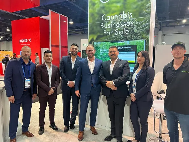 Seven people stand smiling in front of a “Cannabis Businesses For Sale” booth by Green Life Business at a trade show, with red and white display stands and banners highlighting their expertise in cannabis industry business brokerage. Ca NORML