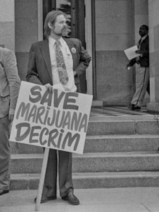 Ca NORML director Dale Gieringer standing with a sign saying "Save Marijuana Decrim" at the state capitol building, sometime in the 70s