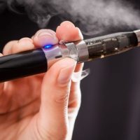 A close-up of a person holding an electronic cigarette, or vape, to their mouth, with vapor visible as they inhale. The device has a blue light and clear chamber showing liquid inside, reflecting progress since 50 years of cannabis reform. CA Norml