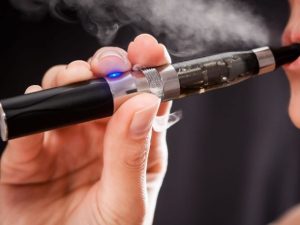 A close-up of a person holding an electronic cigarette, or vape, to their mouth, with vapor visible as they inhale. The device has a blue light and clear chamber showing liquid inside, reflecting progress since 50 years of cannabis reform. CA Norml