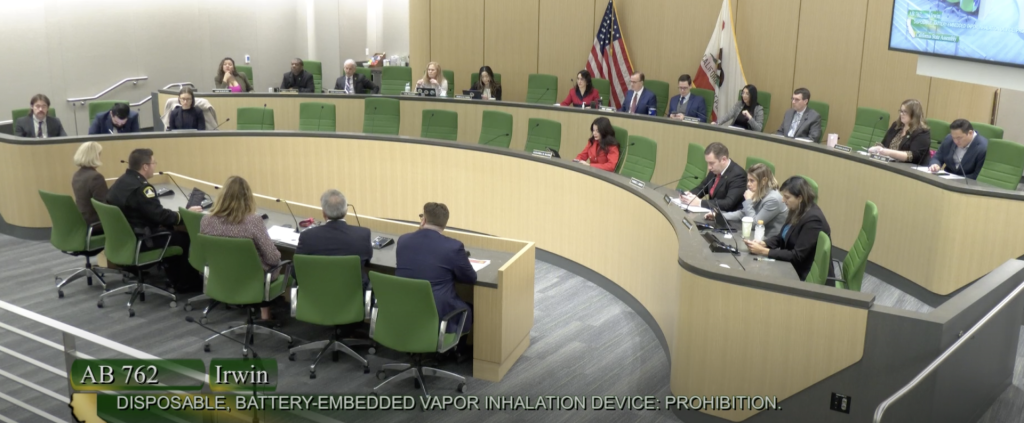 A government committee meets in a large, modern hearing room; people are seated around a curved desk. The screen displays “AB 762 Irwin: Vape ban on Disposable, Battery-Embedded Vapor Inhalation Device: Prohibition.”. CA NORML
